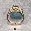 The "Mandevilla" ring in 14k yellow gold with a 1.80-carat Australian sapphire stands upright on rough blue stone against a geometric metallic backdrop, with the Earth’s Treasury logo featured below.