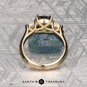 The "Mandevilla" ring in 14k yellow gold with a 1.80-carat Australian sapphire stands upright on rough blue stone against a geometric metallic backdrop, with the Earth’s Treasury logo featured below.