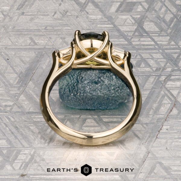 The "Mandevilla" ring in 14k yellow gold with 1.80-carat Australian sapphire The "Mandevilla" ring in 14k yellow gold with a 1.80-carat Australian sapphire stands upright on rough blue stone against a geometric metallic backdrop, with the Earth’s Treasury logo featured below.