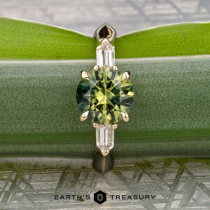 The "Mandevilla" ring in 14k yellow gold, featuring a 1.80-carat Australian sapphire and two rectangular side stones, is shown vertically on a lush green leaf. "EARTH'S TREASURY" appears at the bottom.