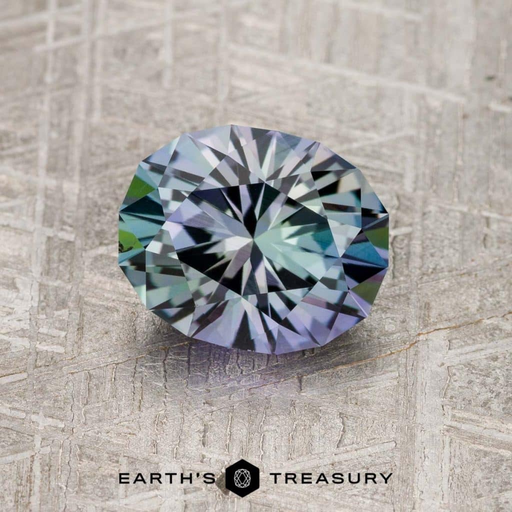 A 4.94-carat Teal-Purple-Green particolored unheated Tanzanite gemstone with a round cut and multifaceted surface, displaying hues of purple and green, laying on a textured surface. The text "EARTH'S TREASURY" is visible at the bottom.