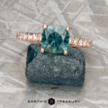 The "Amphitrite" ring in 20K pink gold, featuring a 1.46-carat teal Montana sapphire and diamond accents, rests on a rough blue stone atop a metallic surface. Below, the text "EARTH'S TREASURY" is displayed.