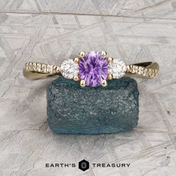 The "Lonicera" ring in 14K yellow gold with 0.66-carat Umba sapphire The "Lonicera" ring in 14K yellow gold features a 0.66-carat round Umba sapphire center, two clear side stones, and small diamonds along the band, presented on a blue rough gemstone.