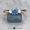 The "Lonicera" ring in 14k rose gold with a 0.93-carat Montana sapphire and two clear stones is displayed on a rough blue mineral, with the Earth's Treasury jewelry logo beneath.
