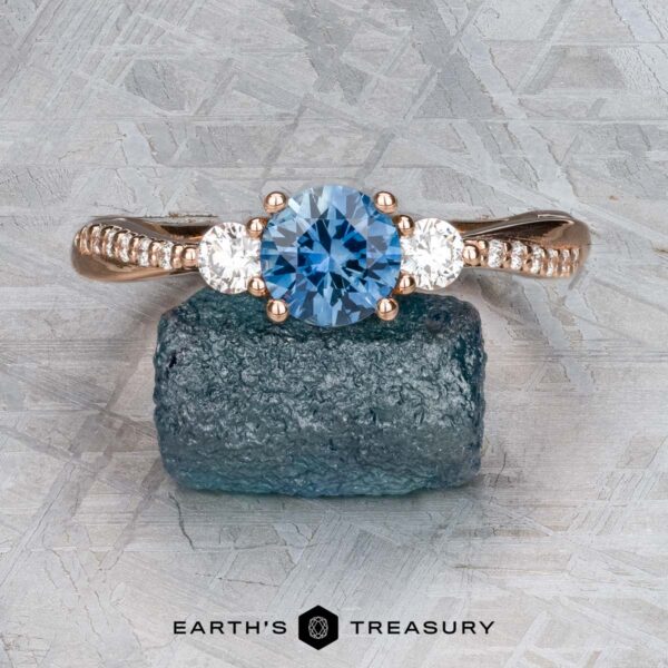 The "Lonicera" ring in 14k rose gold with 0.93-carat Montana sapphire The "Lonicera" ring in 14k rose gold with a 0.93-carat Montana sapphire and two clear stones is displayed on a rough blue mineral, with the Earth's Treasury jewelry logo beneath.