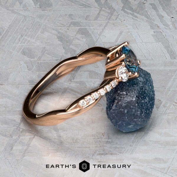 The "Lonicera" ring in 14k rose gold with 0.93-carat Montana sapphire The "Lonicera" ring in 14k rose gold with a 0.93-carat Montana sapphire is displayed beside a rough blue stone on textured gray, with the EARTH'S TREASURY logo below, highlighting nature-inspired jewelry.