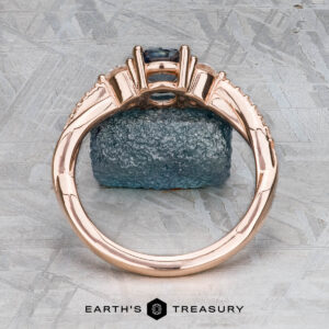 The "Lonicera" ring in 14k rose gold with a 0.93-carat Montana sapphire stands upright on a rough blue stone, set against a gray textured background. The Earth's Treasury jewelry logo appears at the bottom.