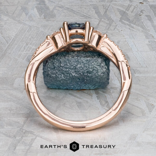 The "Lonicera" ring in 14k rose gold with 0.93-carat Montana sapphire The "Lonicera" ring in 14k rose gold with a 0.93-carat Montana sapphire stands upright on a rough blue stone, set against a gray textured background. The Earth's Treasury jewelry logo appears at the bottom.
