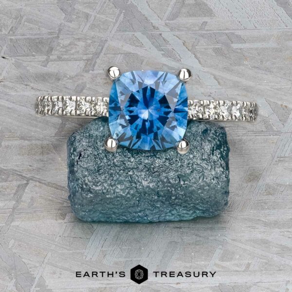 The "Amphitrite" ring in platinum with 1.81-carat Montana sapphire The "Amphitrite" ring in platinum with a 1.81-carat Montana sapphire rests on a rough blue stone atop a gray textured surface, with the "Earth's Treasury" logo appearing at the bottom.