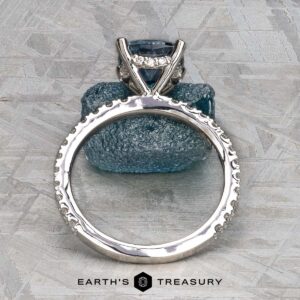 A platinum "Amphitrite" ring with a 1.81-carat Montana sapphire is displayed upright on a textured blue stone background, with the text "EARTH'S TREASURY" below.