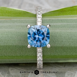 The "Amphitrite" platinum ring, featuring a 1.81-carat Montana sapphire, is displayed upright against a green leaf background.