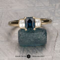 The "Celaeno" ring in 14K yellow gold features a 1.02-carat Australian sapphire and sparkling side stones, elegantly presented on a rough blue mineral with a textured gray background.
