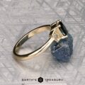 The "Celaeno" ring in 14K yellow gold features a 1.02-carat Australian sapphire and sparkling side stones, elegantly presented on a rough blue mineral with a textured gray background.