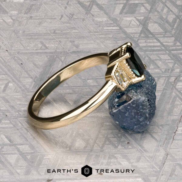 The "Celaeno" ring in 14K yellow gold with 1.02-carat Australian sapphire The "Celaeno" ring in 14K yellow gold features a 1.02-carat Australian sapphire and sparkling side stones, elegantly presented on a rough blue mineral with a textured gray background.