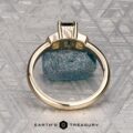 The "Celaeno" ring in 14K yellow gold features a 1.02-carat Australian sapphire and sparkling side stones, elegantly presented on a rough blue mineral with a textured gray background.