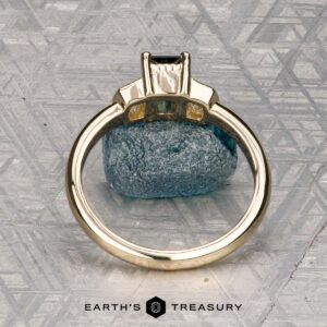 The "Celaeno" ring in 14K yellow gold features a 1.02-carat Australian sapphire and sparkling side stones, elegantly presented on a rough blue mineral with a textured gray background.