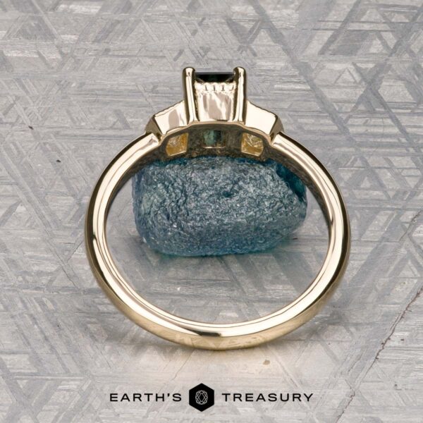 The "Celaeno" ring in 14K yellow gold with 1.02-carat Australian sapphire The "Celaeno" ring in 14K yellow gold features a 1.02-carat Australian sapphire and sparkling side stones, elegantly presented on a rough blue mineral with a textured gray background.