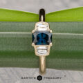 The "Celaeno" ring in 14K yellow gold features a 1.02-carat Australian sapphire and two clear geometric stones, elegantly displayed on a green leaf with The Earth's Treasury logo at the bottom.