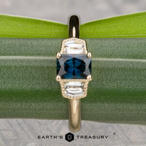 The "Celaeno" ring in 14K yellow gold features a 1.02-carat Australian sapphire and two clear geometric stones, elegantly displayed on a green leaf with The Earth's Treasury logo at the bottom.