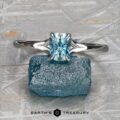 The "Cybele" ring in 18K white gold, featuring a 0.96-carat rectangular Montana sapphire, is showcased atop a rough blue mineral against a textured gray background—an elegant design inspired by Cybele.
