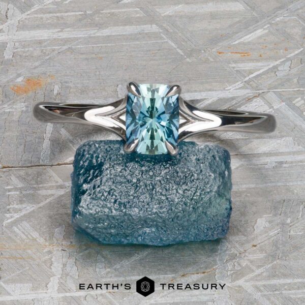 The "Cybele" ring in 18K white gold with 0.96-carat Montana sapphire The "Cybele" ring in 18K white gold, featuring a 0.96-carat rectangular Montana sapphire, is showcased atop a rough blue mineral against a textured gray background—an elegant design inspired by Cybele.