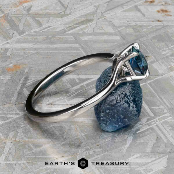 The "Cybele" ring in 18K white gold with 0.96-carat Montana sapphire The "Cybele" ring in 18K white gold featuring a 0.96-carat Montana sapphire is displayed atop a raw blue stone on a textured gray surface. The Earth's Treasury logo appears below, capturing Cybele's elegance.