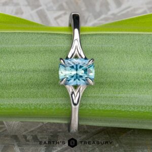 An 18K white gold "Cybele" solitaire engagement ring with a 0.96-carat Montana sapphire displayed on a green leaf, with the Earth's Treasury logo at the bottom.