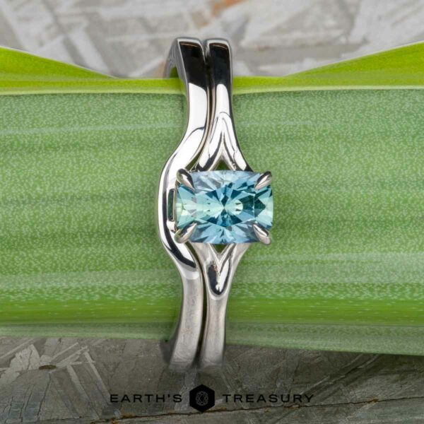 The "Cybele" ring in 18K white gold with 0.96-carat Montana sapphire, alongside the custom-fit contour band in 18K white gold The "Cybele" ring in 18K white gold with a 0.96-carat Montana sapphire and its custom-fit contour band rest on a green leaf, with the "Earth's Treasury" logo at the bottom.