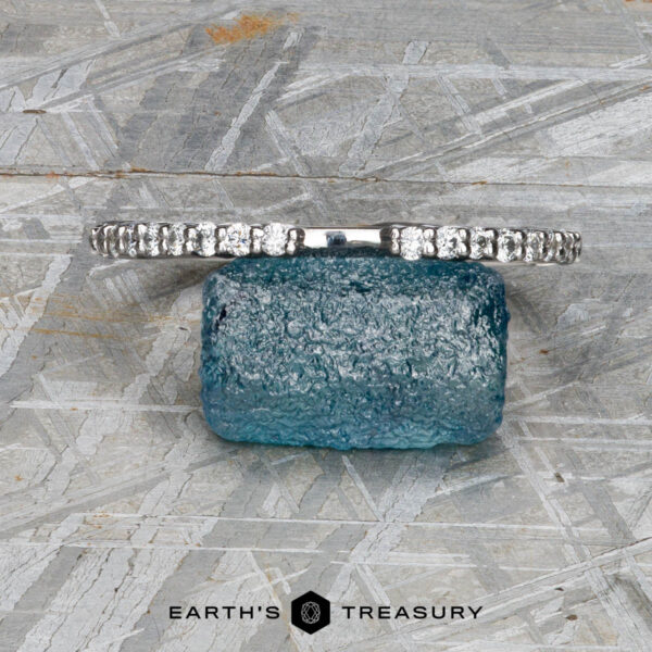 The "Kivu" band in 18k white gold The "Kivu" band in 18k white gold, designed in a Hidden Halo Solitaire style, rests on a rough blue gemstone against a textured gray background, with the Earth's Treasury logo below.