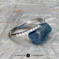 The "Kivu" band in 18k white gold, set with small diamonds in a Hidden Halo Solitaire style, rests beside a rough blue gemstone on a metallic textured surface, with the Earth’s Treasury logo below.