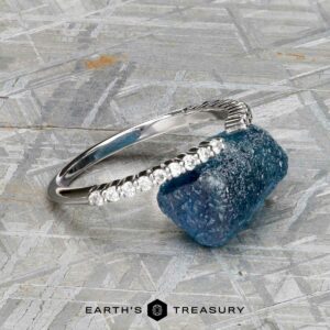 The "Kivu" band in 18k white gold, set with small diamonds in a Hidden Halo Solitaire style, rests beside a rough blue gemstone on a metallic textured surface, with the Earth’s Treasury logo below.