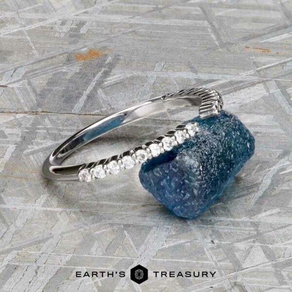 The "Kivu" band in 18k white gold The "Kivu" band in 18k white gold, set with small diamonds in a Hidden Halo Solitaire style, rests beside a rough blue gemstone on a metallic textured surface, with the Earth’s Treasury logo below.