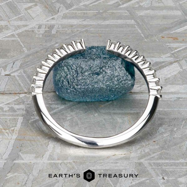 The "Kivu" band in 18k white gold The "Kivu" band in 18k white gold stands upright before a rough blue stone on a gray, patterned surface. "Earth's Treasury" is displayed at the bottom.