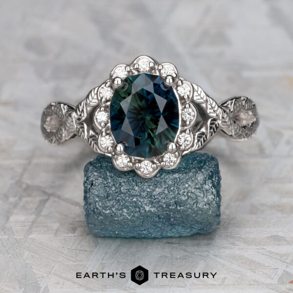 The "Passiflora" ring in 14k white gold with 2.48-carat Australian sapphire The "Passiflora" ring in 14k white gold features a 2.48-carat Australian sapphire, accented with small clear stones and displayed on a blue mineral, with the Earth’s Treasury logo shown below.