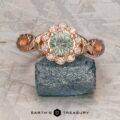 The "Passiflora" ring in 14K rose gold features a 0.93-carat round Montana sapphire center with a diamond halo, elegantly displayed on a rough blue stone.
