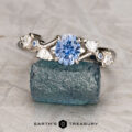 The "Jasmine" ring in 14K white gold, featuring a 0.91-carat Montana sapphire and four accent diamonds, is displayed on a rough blue-green mineral with "Earth's Treasury" at the bottom.