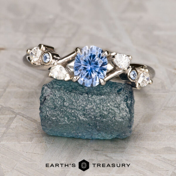 The "Jasmine" ring in 14K white gold with 0.91-carat Montana sapphire The "Jasmine" ring in 14K white gold, featuring a 0.91-carat Montana sapphire and four accent diamonds, is displayed on a rough blue-green mineral with "Earth's Treasury" at the bottom.