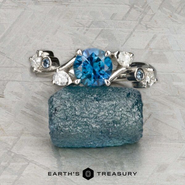The "Jasmine" ring in platinum with 1.05-carat Montana sapphire A platinum "Jasmine" ring with a 1.05-carat Montana sapphire centerpiece, accented by diamonds, rests on a rough blue stone. The "EARTH'S TREASURY" logo and text are displayed at the bottom.