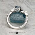 The "Jasmine" ring in platinum with a 1.05-carat Montana sapphire rests on a rough blue stone atop a textured gray surface, with the Earth's Treasury logo below—an elegant jewelry piece for any occasion.