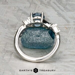 The "Jasmine" ring in platinum with a 1.05-carat Montana sapphire rests on a rough blue stone atop a textured gray surface, with the Earth's Treasury logo below—an elegant jewelry piece for any occasion.