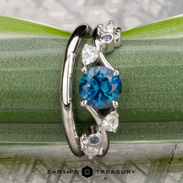 The "Jasmine" ring in platinum with 1.05-carat Montana sapphire, alongside the custom-fit contour band in platinum The "Jasmine" ring in platinum, featuring a 1.05-carat Montana sapphire and custom-fit contour band, is displayed on a green leaf. The "Earth's Treasury" logo appears below, highlighting this elegant gemstone ring set.