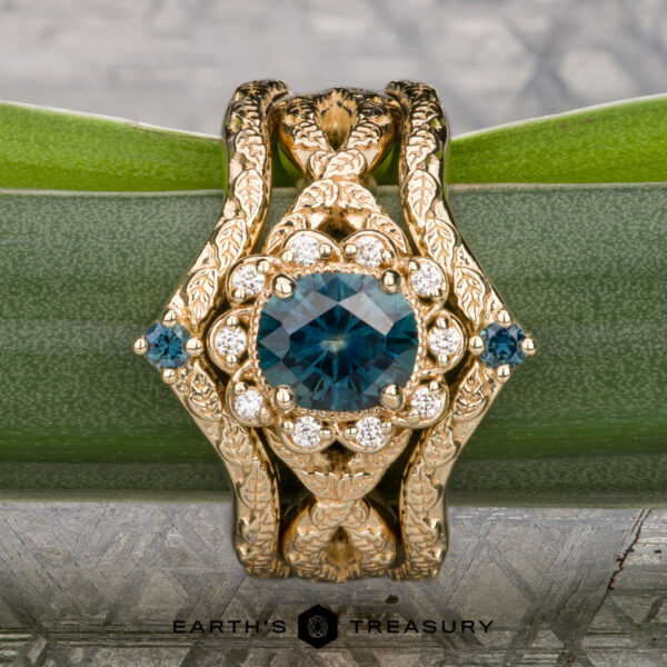 The "Passiflora" ring in 14K yellow gold with 1.10-carat Montana sapphire, alongside two "Passiflora" bands in 14K yellow gold The "Passiflora" ring in 14K yellow gold with a 1.10-carat Montana sapphire is shown with two matching "Passiflora" bands, all crafted in 14K yellow gold and elegantly displayed on a green stem.
