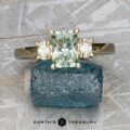 The "Dryope" ring in 14k yellow gold features a 1.56-carat rectangular pale green Montana sapphire flanked by two clear stones, displayed on a rough blue mineral with the "Earth's Treasury" logo below.