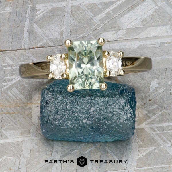 The "Dryope" ring in 14k yellow gold with 1.56-carat Montana sapphire The "Dryope" ring in 14k yellow gold features a 1.56-carat rectangular pale green Montana sapphire flanked by two clear stones, displayed on a rough blue mineral with the "Earth's Treasury" logo below.