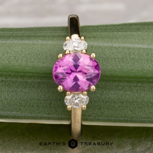 The "Dryope" ring in 18k yellow gold features a 1.41-carat Ceylon sapphire between two clear stones, elegantly displayed on a green textured surface with "Earth's Treasury" below.