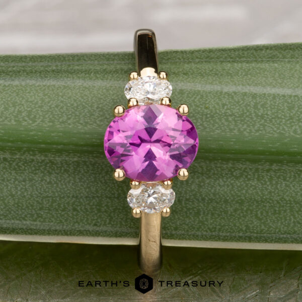 The "Dryope" ring in 18k yellow gold with 1.41-Carat Ceylon sapphire The "Dryope" ring in 18k yellow gold features a 1.41-carat Ceylon sapphire between two clear stones, elegantly displayed on a green textured surface with "Earth's Treasury" below.