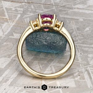 The "Dryope" ring in 18k yellow gold showcases a 1.41-carat Ceylon sapphire flanked by two diamonds, displayed upright on textured blue stone with the Earth's Treasury logo below. Inspired by the enchanting Dryope collection.
