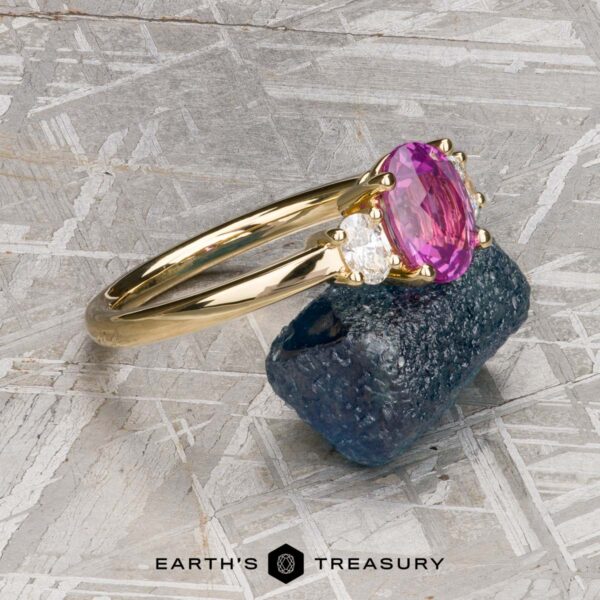 The "Dryope" ring in 18k yellow gold with 1.41-Carat Ceylon sapphire The "Dryope" ring in 18k yellow gold, featuring a 1.41-carat Ceylon sapphire and two side diamonds, is displayed on a rough blue stone with Earth's Treasury branding at the bottom.