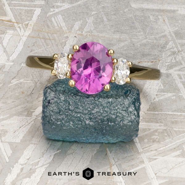 The "Dryope" ring in 18k yellow gold with 1.41-Carat Ceylon sapphire The "Dryope" ring in 18k yellow gold features a 1.41-carat Ceylon sapphire accented by two white stones, displayed on a blue-green mineral with Earth's Treasury branding below. Inspired by Dryope, this three-stone piece is truly unique.
