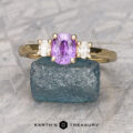 The "Dryope" ring features a 1.03-carat Madagascar sapphire set in 14K yellow gold, flanked by two clear stones and displayed on a rough blue crystal with "Earth's Treasury" beneath.